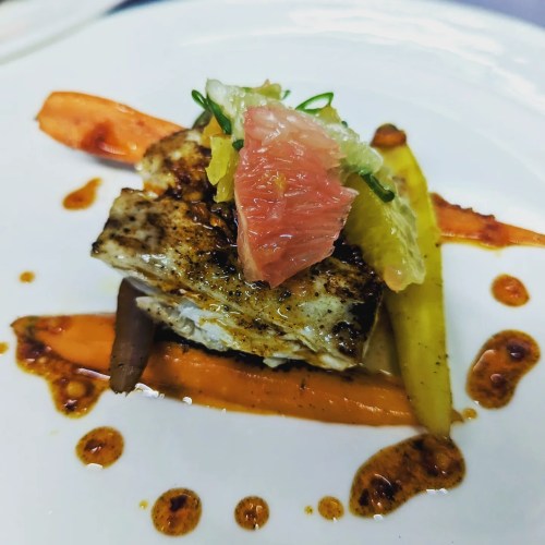 A beautifully plated dish featuring grilled fish, colorful vegetables, and citrus garnishes on a white plate.