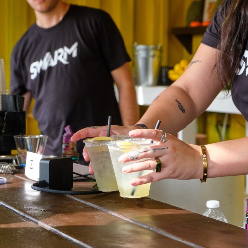 Two glasses of lemonade with lemon slices on a wooden bar, with a person preparing drinks in the background.