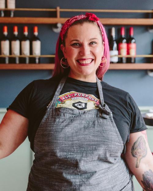 A smiling chef with pink hair and a bandana wears a gray apron over a black shirt, standing in a restaurant.