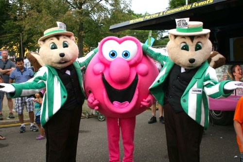Two mascots in green jackets stand beside a large pink character, all smiling at a fair with people in the background.