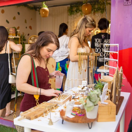 A woman browses jewelry at a colorful market stall while another shopper looks on in the background.