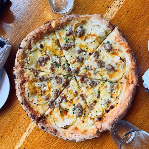 A round pizza topped with sausage, cheese, and herbs, served on a wooden table with glasses nearby.