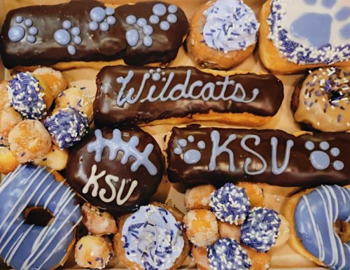 A box of decorated donuts featuring blue and chocolate icing, with designs including "Wildcats" and "KSU."