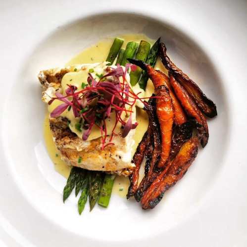 A plated dish featuring fish on asparagus, topped with sauce and microgreens, alongside roasted carrots.