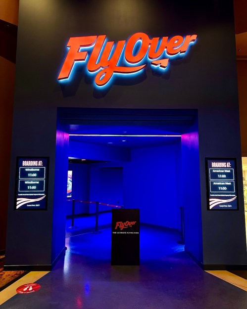 Entrance to "FlyOver" attraction, featuring blue lighting and digital boards displaying boarding times.
