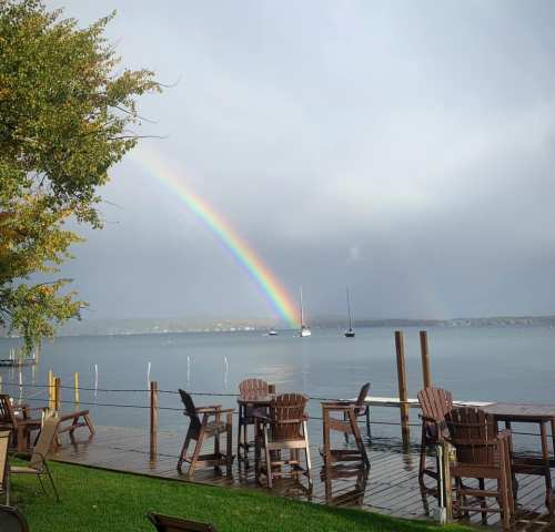 A vibrant rainbow arcs over a calm lake, with boats in the distance and wooden chairs along the shore.