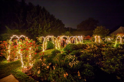 A beautifully lit garden at night, featuring archways adorned with string lights and vibrant flowers.
