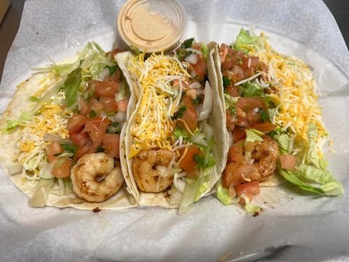 Three shrimp tacos topped with lettuce, tomatoes, and cheese, served with a side of sauce in a small container.