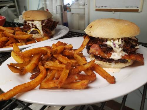 A close-up of a burger with pulled pork, bacon, and coleslaw, served with crispy sweet potato fries on a white plate.