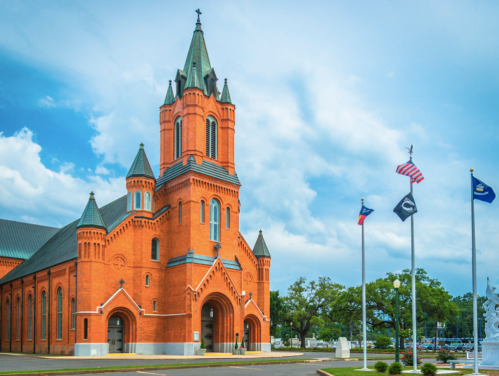 Historic red brick church with a tall steeple, surrounded by flags and a blue sky with scattered clouds.