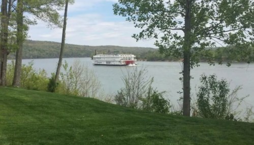 A riverboat glides along a calm river, surrounded by lush greenery and rolling hills under a partly cloudy sky.