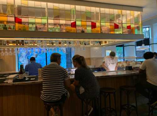 A modern bar with colorful glass decor, featuring patrons seated and a chef preparing food in the background.