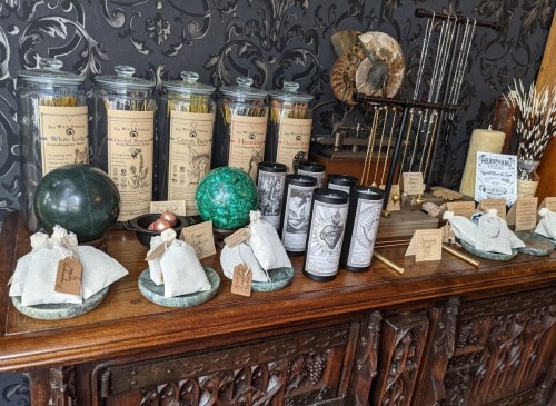 A wooden table displays jars of herbs, candles, and decorative items against a dark patterned background.