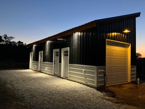 A modern metal building with white doors, illuminated at dusk, surrounded by gravel and trees in the background.