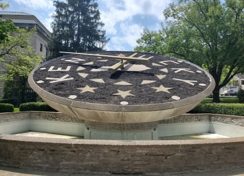 A large, circular sundial with "Kentucky" and stars, surrounded by greenery and a stone wall.