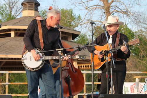 Two musicians perform on stage, one playing a banjo and the other an acoustic guitar, with a double bass in the background.