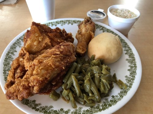 A plate of fried chicken, green beans, a roll, and a cup of coleslaw on a wooden table.