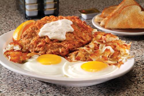A plate of fried chicken with gravy, two sunny-side-up eggs, hash browns, and toast, garnished with orange slices.