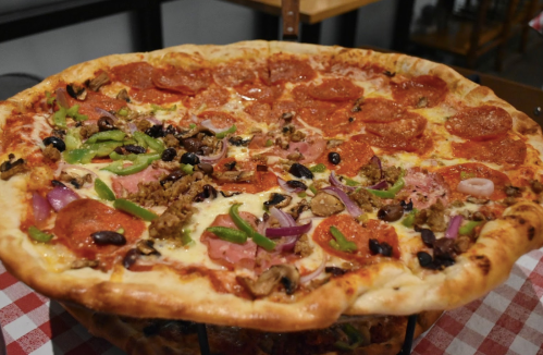 A large pizza with half pepperoni and half assorted toppings, including green peppers, olives, and mushrooms, on a checkered tablecloth.