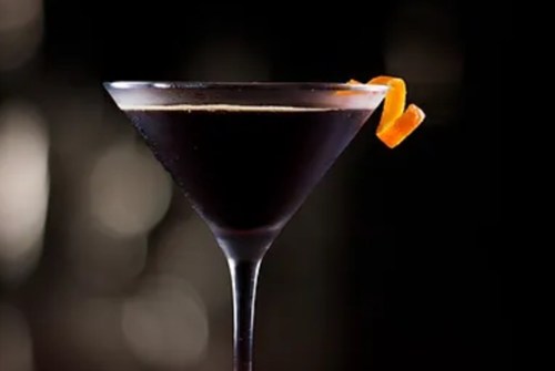 A dark cocktail in a martini glass, garnished with a twist of orange peel, set against a blurred background.