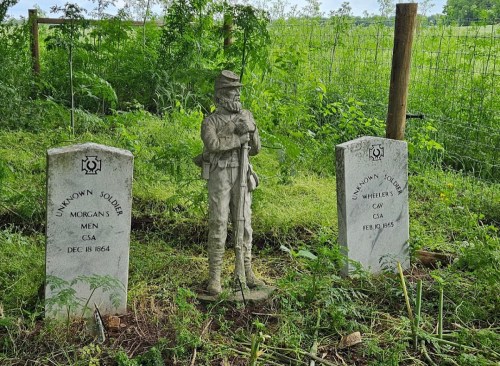 Two gravestones marked "Unknown Soldier" flanking a soldier statue, set in a grassy area with a wooden fence in the background.