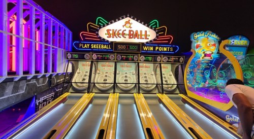 A brightly lit skee ball arcade game with three lanes and colorful neon signage in the background.