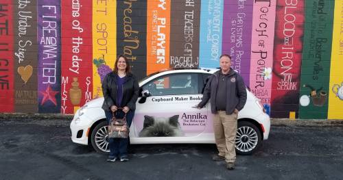A woman and a man stand beside a car featuring a cat graphic and the name "Annika, the Bookstore Cat," in front of a colorful mural.