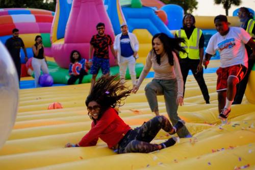 Two women playfully slide on a colorful inflatable surface, surrounded by people enjoying a fun outdoor event.