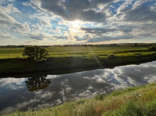 A serene river reflects a cloudy sky and sun, surrounded by lush green fields and trees.