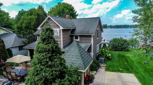 A charming house with a sloped roof, surrounded by greenery, overlooking a serene lake under a blue sky.
