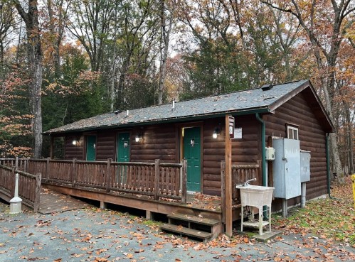 A rustic wooden cabin with green doors, surrounded by autumn foliage and a wooden deck.