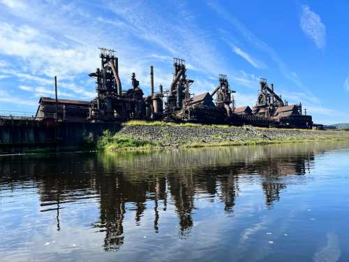 Industrial structures along a river, reflecting in the water under a clear blue sky.