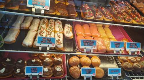 A display case filled with various long john donuts, including chocolate, vanilla, strawberry, and more, with colorful toppings.