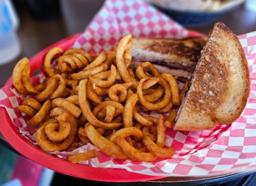A plate with curly fries and a grilled sandwich on toasted bread, served in a red basket with checkered paper.