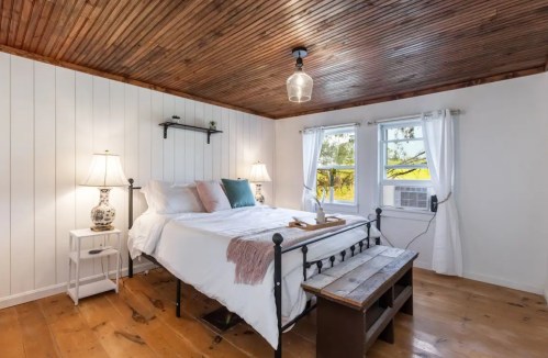 Cozy bedroom with a wooden ceiling, a bed, two lamps, and a bench by the window, featuring natural light and a serene atmosphere.