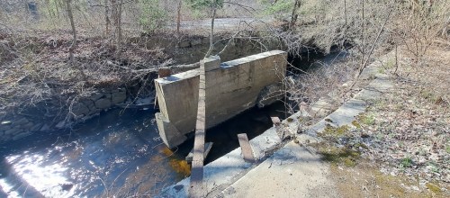 A concrete structure partially submerged in a shallow stream, surrounded by bare trees and rocky terrain.