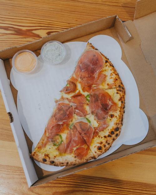 A slice of pizza topped with prosciutto, served in a takeout box with two dipping sauces on the side.