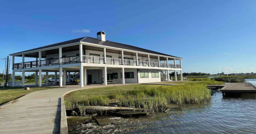 A modern two-story building by the water, surrounded by grass and a wooden walkway leading to a dock.