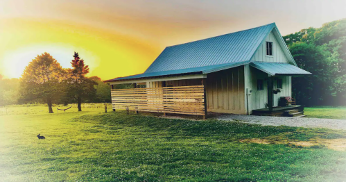 A cozy house with a metal roof, surrounded by green grass and trees, under a vibrant sunset. A rabbit is nearby.