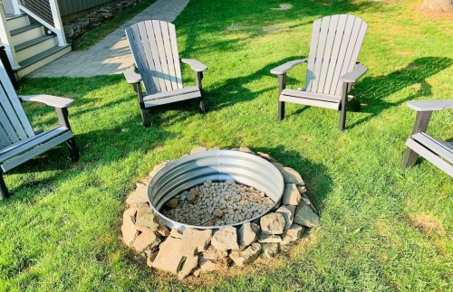 A circular fire pit surrounded by stone, with four gray Adirondack chairs on a grassy lawn.