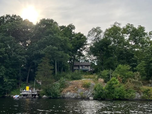 A serene lakeside view featuring a house on a hill, surrounded by trees, with a small dock and boat in the foreground.