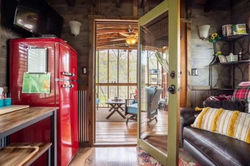 Cozy cabin interior with a red fridge, wooden decor, and a view of a porch with seating and greenery outside.