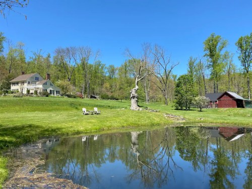 A serene landscape featuring a house, a pond reflecting trees, and a red barn under a clear blue sky.