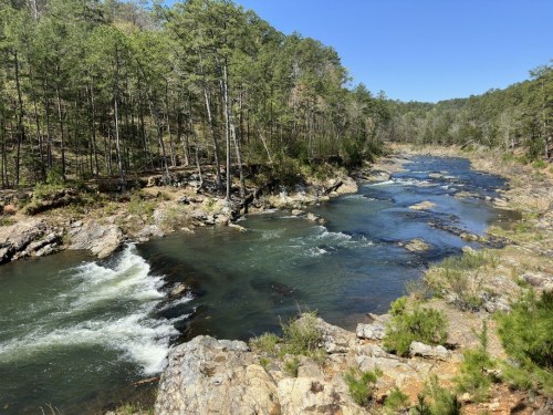 A serene river flows through a rocky landscape, surrounded by lush green trees under a clear blue sky.