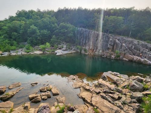 A serene rocky landscape with a calm greenish pool surrounded by lush trees and cliffs under a clear sky.