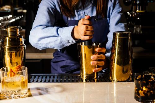 A bartender in a blue shirt mixes a cocktail using a gold shaker at a stylish bar.