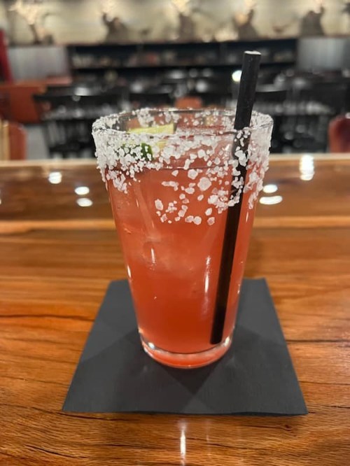 A cocktail with a salted rim, garnished with a lime slice, sits on a black coaster on a wooden bar.
