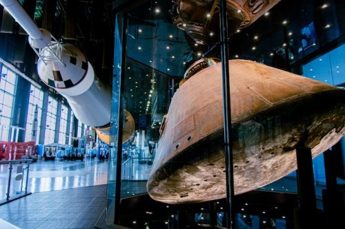 A close-up of a spacecraft capsule displayed in a modern museum, with a rocket in the background and bright lighting.