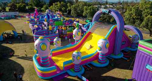 A colorful inflatable slide with cartoonish designs, surrounded by various inflatable attractions in a park setting.