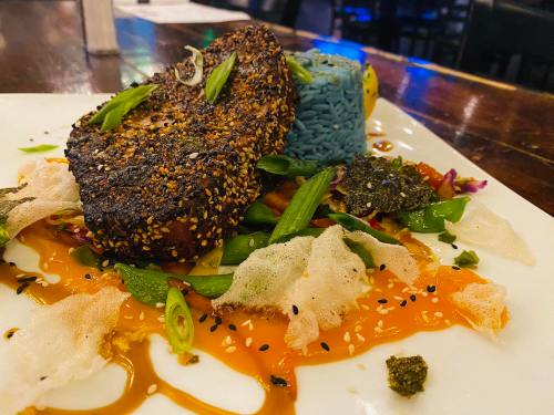 A plate featuring a seasoned fish fillet, blue rice, colorful vegetables, and sauces, garnished with green onions.